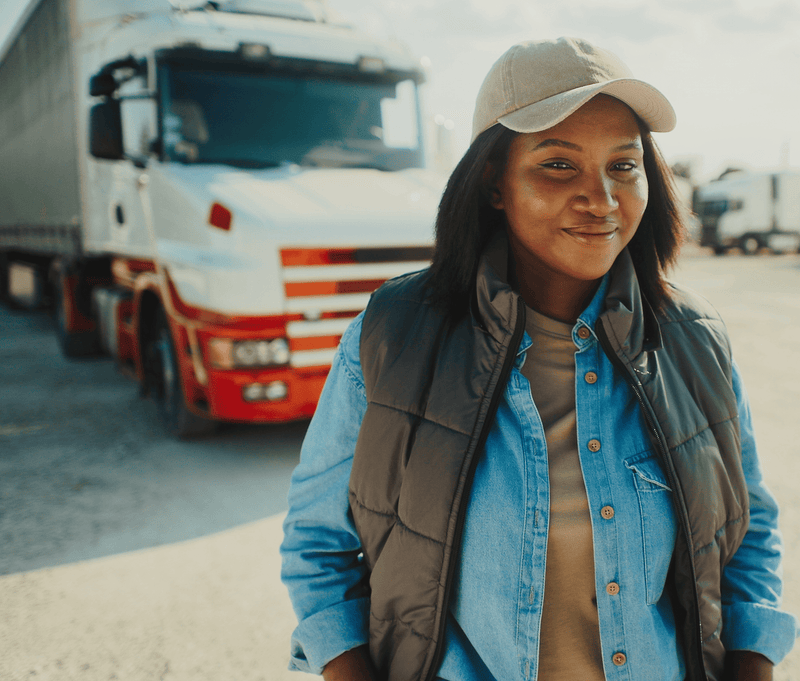 Young woman in a denim shirt and cap smiles confidently, standing in front of a large truck.