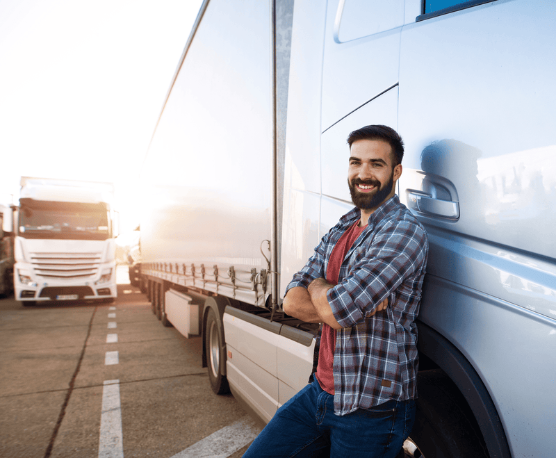 Man leaning against a large blue truck, smiling confidently. Dressed in a plaid shirt, with arms crossed. Bright day, with another truck in the background.