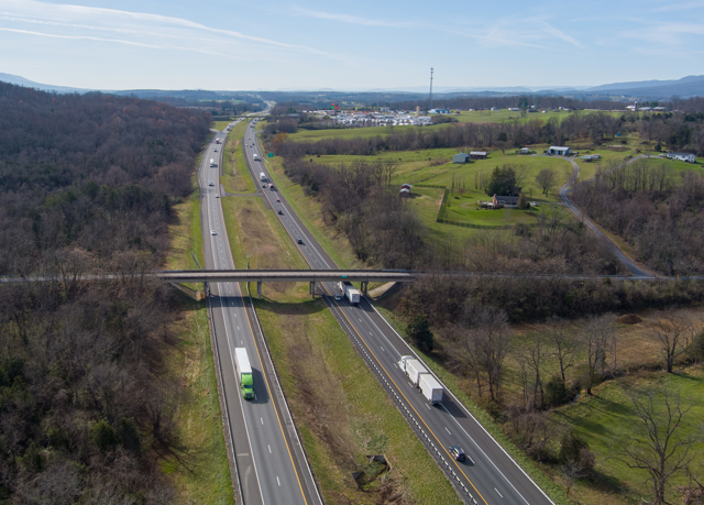 Aerial view of a highway with light traffic, including trucks and cars, surrounded by green fields and sparse trees. Clear sky and distant hills convey a calm, expansive scene.