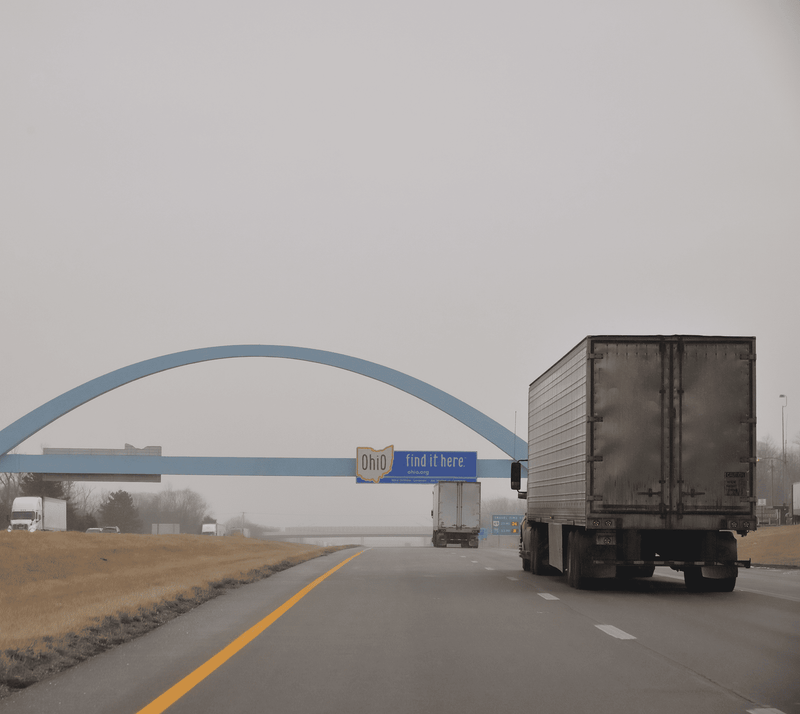 A gray semi-truck drives on an empty highway under a blue archway in Ohio