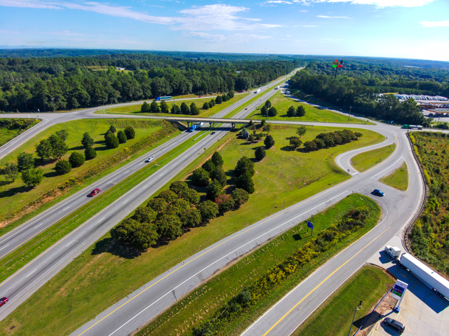 Aerial view of a highway interchange with curving roads, greenery, and scattered trees under a clear blue sky, conveying a sense of openness.